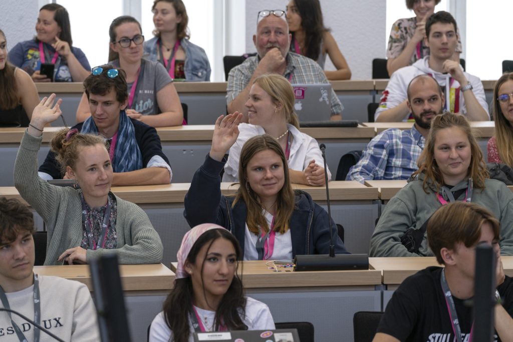 A group of people are sitting in rows and listening to someone outside of the frame of the picture. Two young women raise their hands.