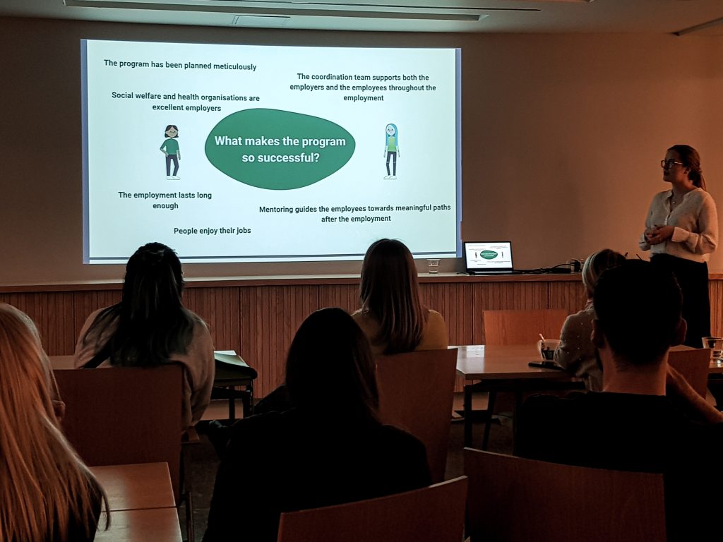 People are watching a Powerpoint presentation. The text says: "What makes the program so successful? The program has been planned meticulously. The coordination team supports both the employers and the employee throughout the employment. The employment lasts long enough. People enjoy their jobs. Mentoring guides the employees towards meaningful employment."